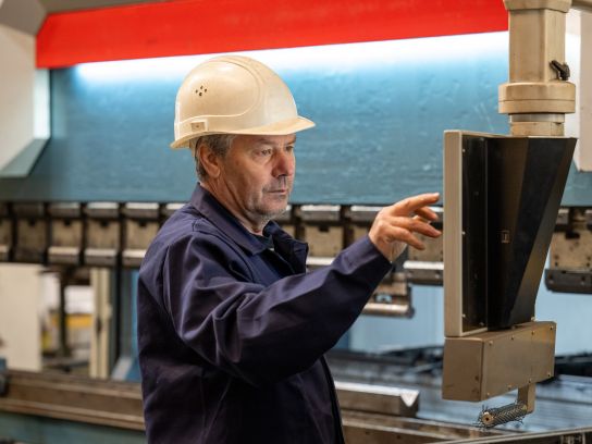 Stapu-Mitarbeiter bei der Programmierung einer CNC gesteuerten Anlage in der Fertigungshalle. Das Bild zeigt einen der Arbeitsschritte innerhalb der mechanischen Anarbeitungsverfahren wie Fräsen oder Gewindeschneiden.