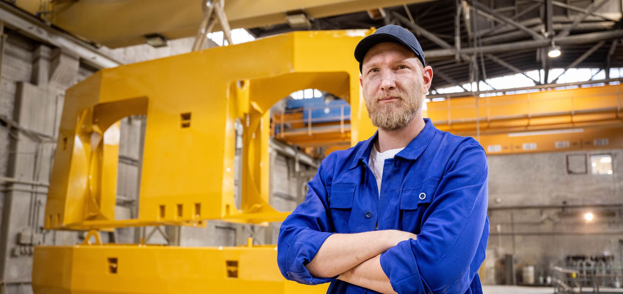 Ein Mitarbeiter der Stapu GmbH & Co. KG in blauer Arbeitskleidung steht mit verschränkten Armen vor einem massiven, gelben Gegengewicht in der Fertigungshalle.