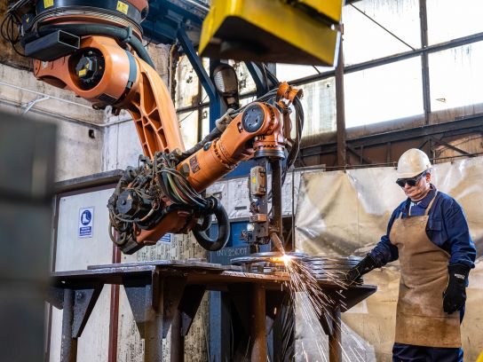 Ein leistungsstarker Industrie-Roboter beim automatisierten Autogen-Fasenschneiden von dicken Stahlblechen. Ein Mitarbeiter in Schutzkleidung überwacht den Prozess der maßgenauen Brennschnitte in der Werkshalle.