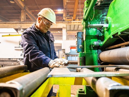 Eine schwere Industriemaschine biegt eine massive Stahlplatte in der Fertigungshalle von Stapu. Der Fokus liegt auf der präzisen Verformung des dicken Materials und der enormen Kraft, die bei diesem Umformprozess zum Einsatz kommt.