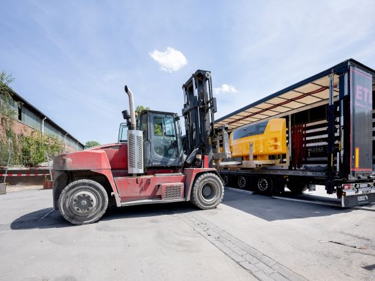 Verladung eines fertig bearbeiteten und lackierten Gegengewichts auf dem Werksgelände. Die Aufnahme dokumentiert die Handhabung schwerer Lasten im Rahmen der Logistikkette.