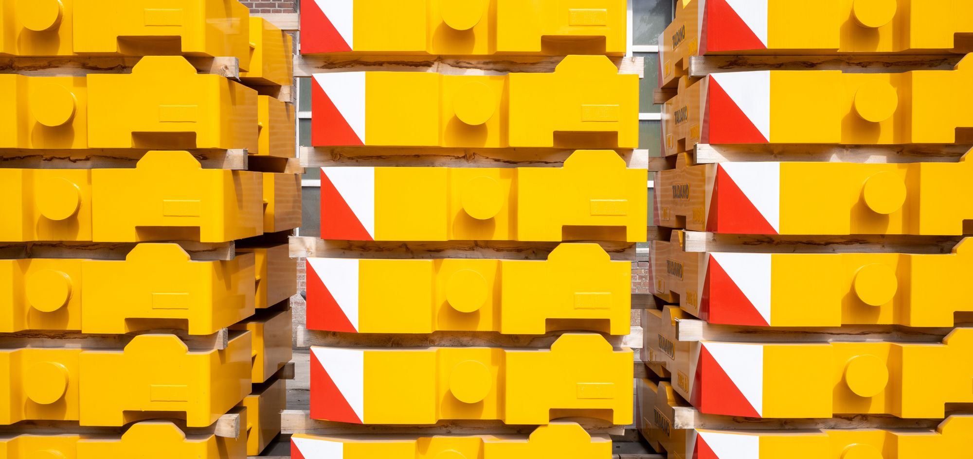 Mehrere Stapel fertig lackierter gelber Gegengewichte lagern versandbereit auf dem Betriebsgelände. Die markanten Bauteile sind mit rot weißen Warnmarkierungen versehen und lagern sicher auf Holzunterlagen für den weiteren Transport.