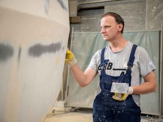 Ein Mitarbeiter der Stapu GmbH & Co. KG in Schutzkleidung trägt eine Grundierung auf ein massives Stahlbauteil auf. Die gleichmäßige Farbschicht schützt das Material vor Korrosion und bereitet es für den industriellen Einsatz vor.