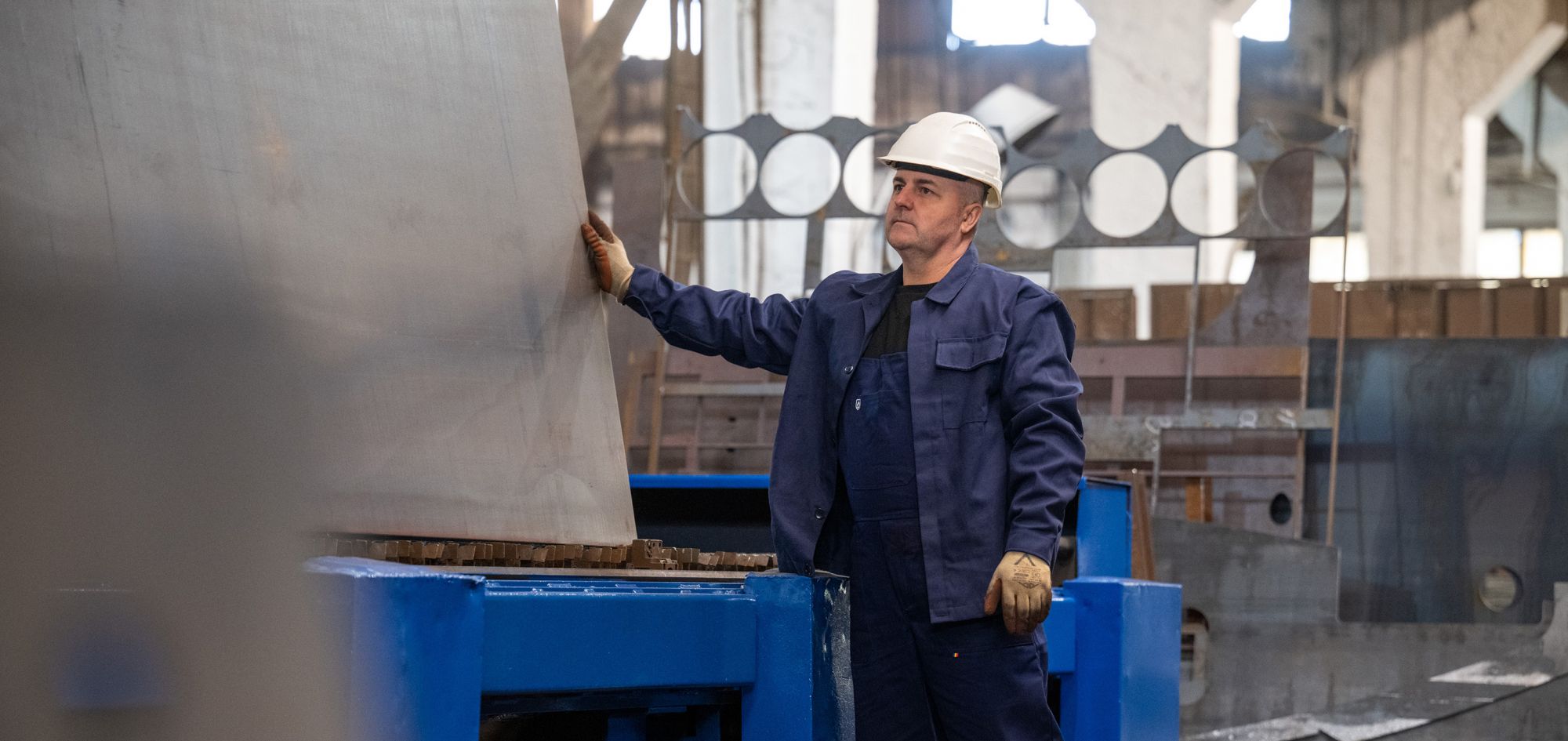 Stapu-Fachkraft im Stahlbau bei der Arbeit mit massiven Metallbauteilen in einer Fertigungshalle. Der Arbeiter trägt Schutzausrüstung und begutachtet eine vertikal ausgerichtete Stahlplatte.